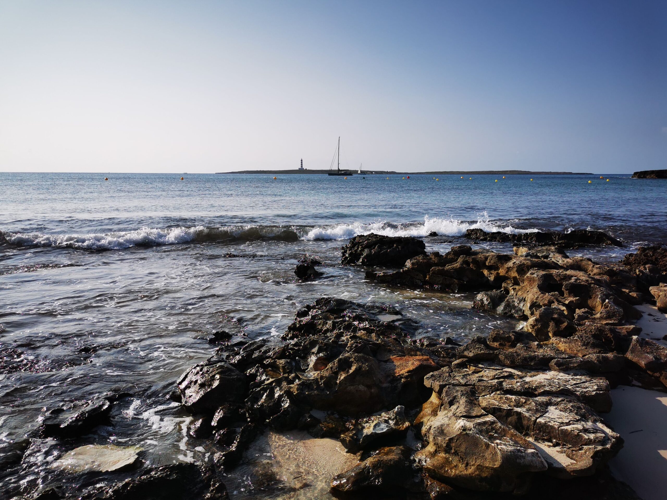 Serene seascape at Punta Prima, Spain, featuring a rocky shoreline and distant sailboat under clear skies.